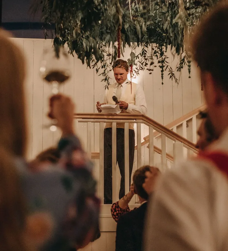 Man giving a speech from a balcony while guests listen and raise glasses at an indoor celebration