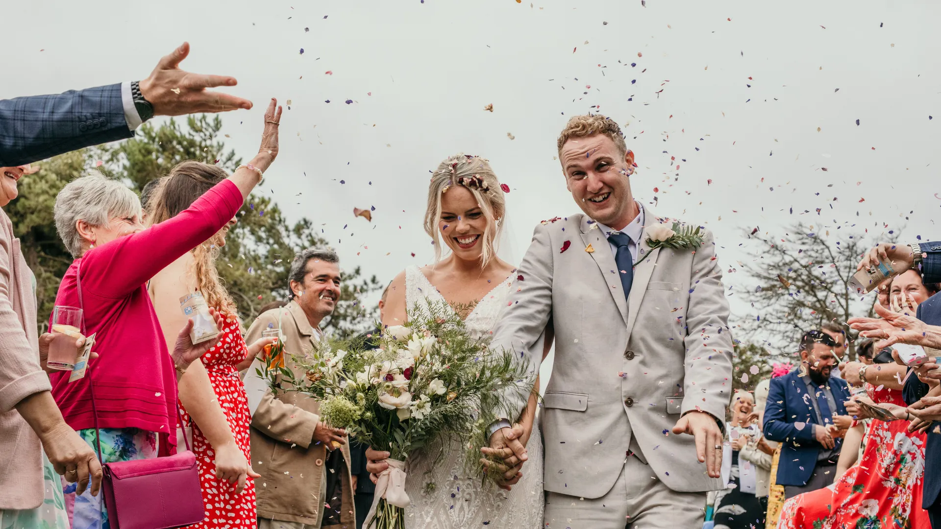 Bride and groom walk through guests throwing colourful confetti at an outdoor wedding