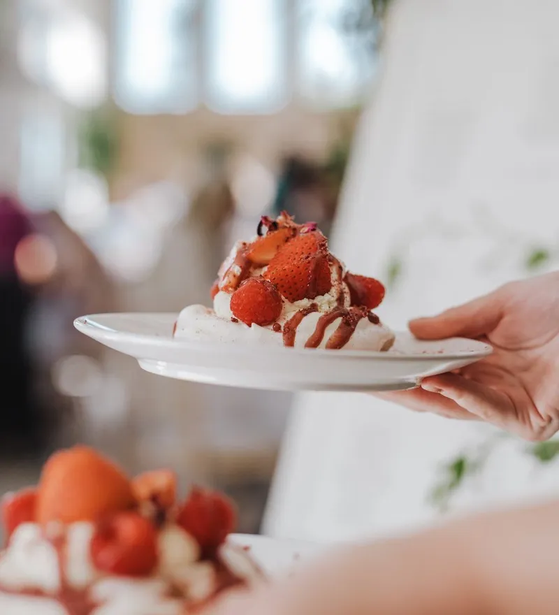 Hand holding a white plate with a strawberry and cream dessert drizzled with chocolate in a bright café