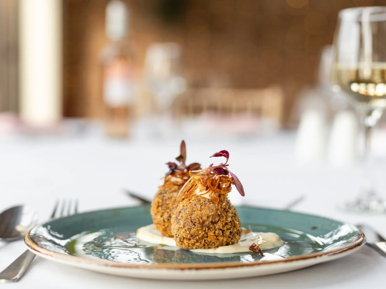 Two crumbed croquettes served on a blue-green plate with a creamy sauce and microgreens, with a glass of white wine in the background.