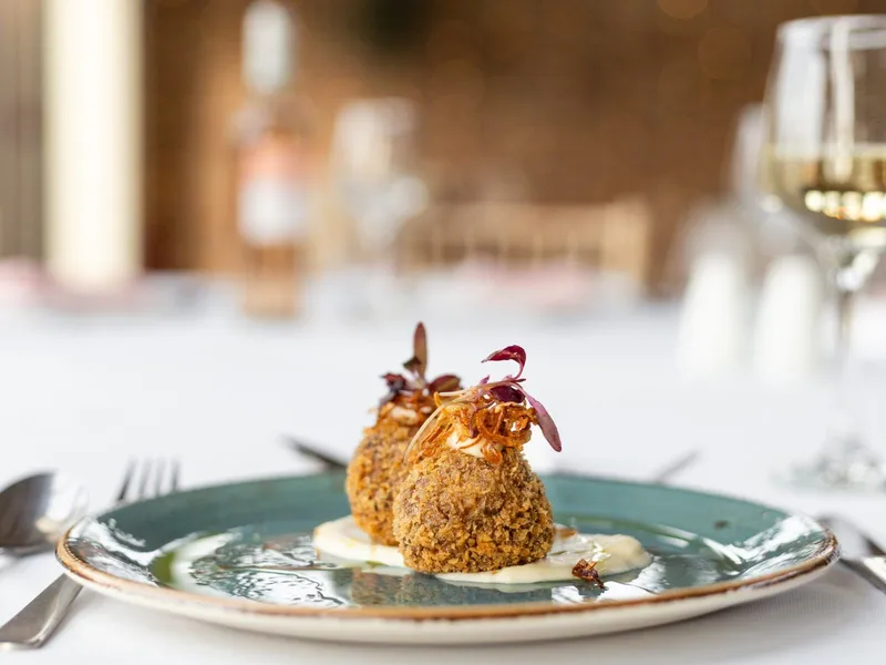 Two crumbed croquettes served on a blue-green plate with a creamy sauce and microgreens, with a glass of white wine in the background.