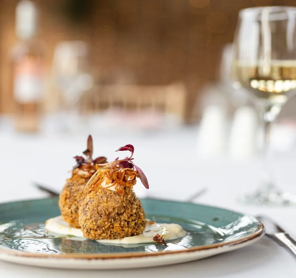 Two breadcrumb-coated croquettes on a blue-green plate with sauce and microgreens, with a glass of white wine in the background