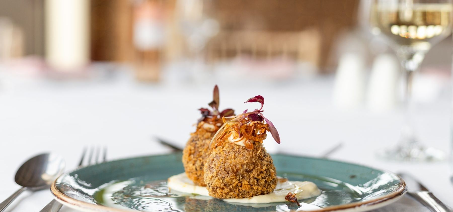 Two breadcrumb-coated croquettes on a blue-green plate with sauce and microgreens, with a glass of white wine in the background