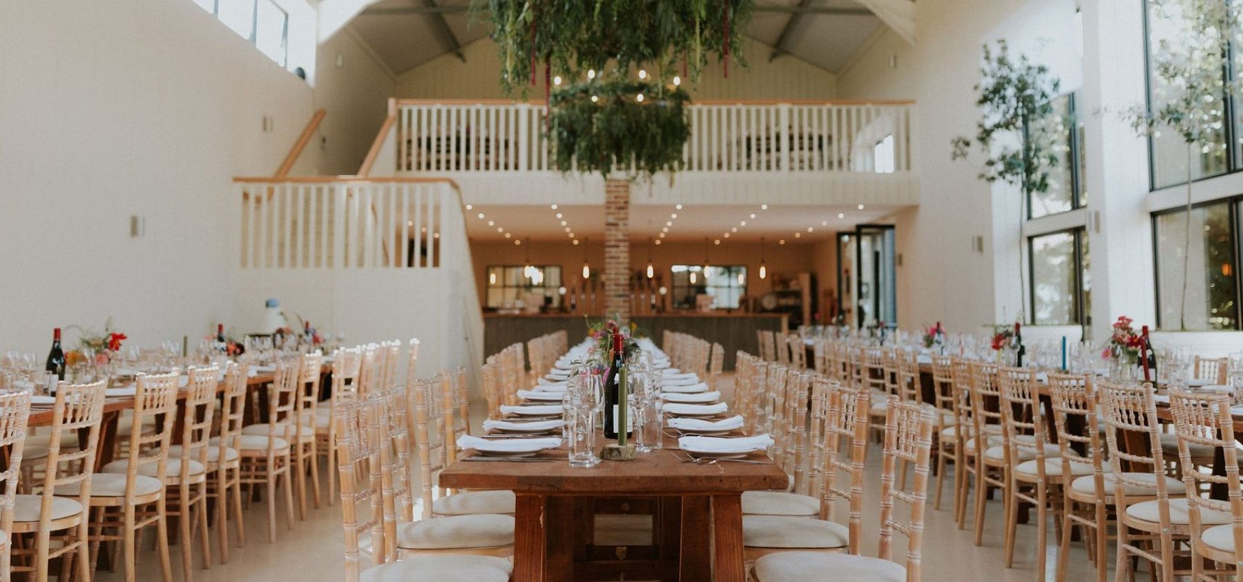 Long banquet tables set for a wedding reception in a bright white barn with a large greenery chandelier overhead
