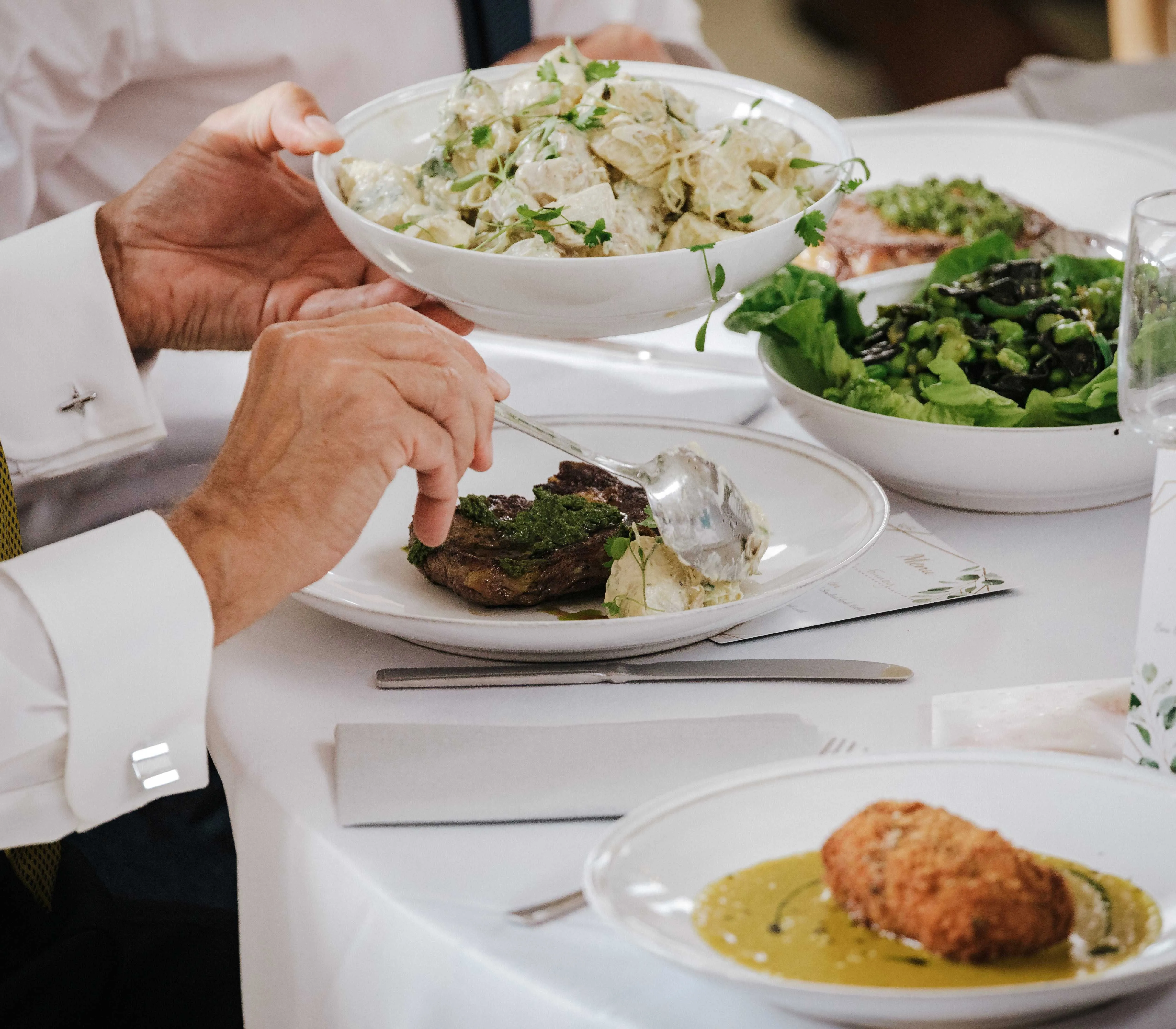 Person serving potato salad at a formal dinner table with steak and green salad
