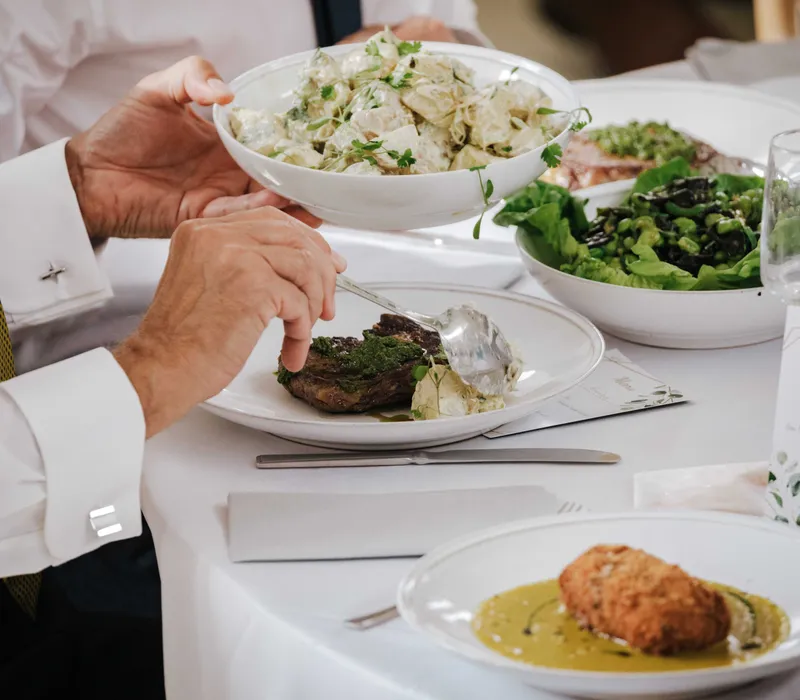 Person serving potato salad at a formal dinner table with steak and green salad