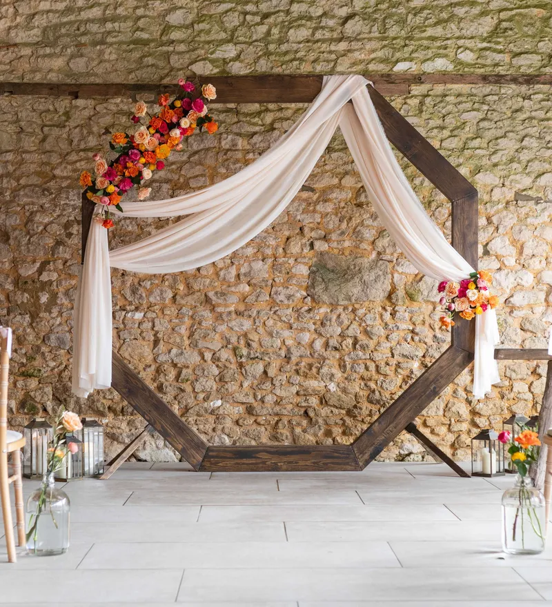 Rustic wedding ceremony setup with hexagonal wooden arch draped in white fabric and colourful flowers against a stone wall