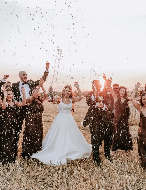 Wedding party celebrating in a field at sunset as champagne sprays, with the bride in a white gown and guests cheering