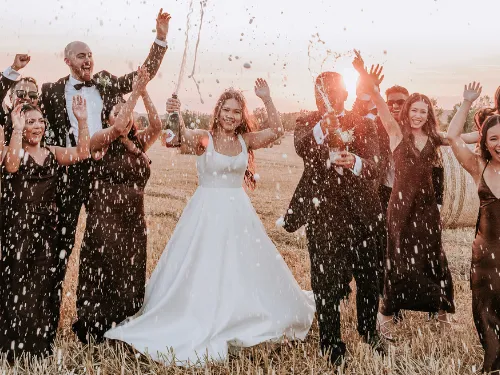 Wedding party celebrating in a field at sunset as champagne sprays, with the bride in a white gown and guests cheering