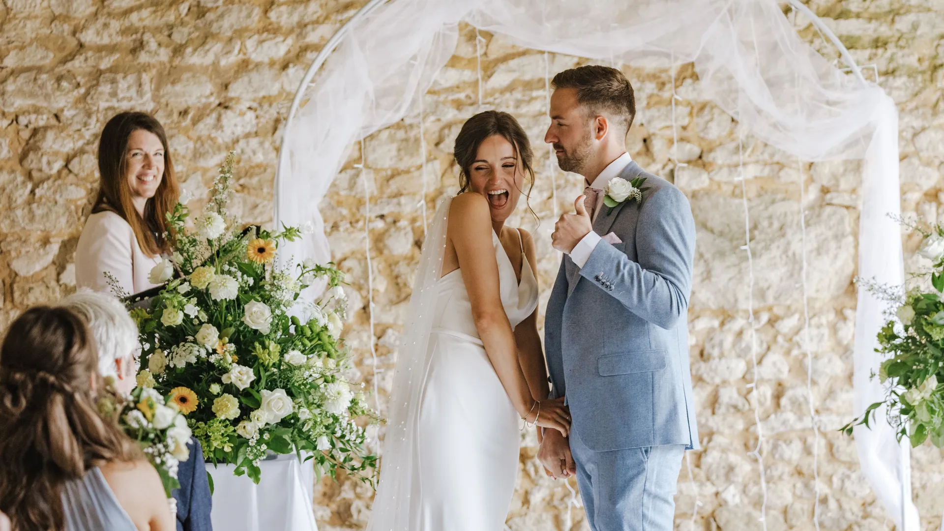 Bride laughing beside groom under a white draped wedding arch, with an officiant and flowers in front of a stone wall.