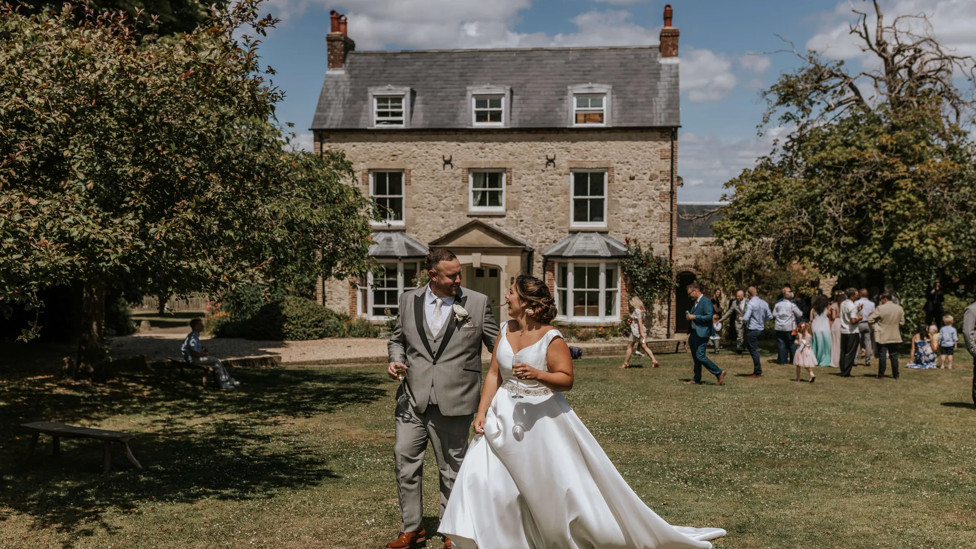 Bride and groom walking on a lawn in front of a stone country house during an outdoor wedding reception