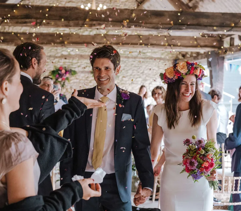 Newlyweds walk through cheering guests as confetti falls indoors, with the bride wearing a floral crown and holding a colourful bouquet.