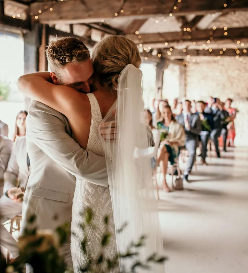 Bride and groom embrace during a rustic indoor wedding ceremony with fairy lights overhead