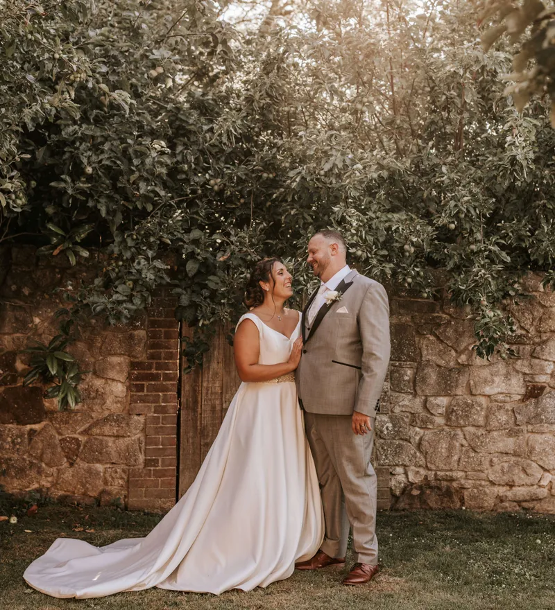 Bride and groom smiling together in a garden beside a stone wall beneath leafy trees