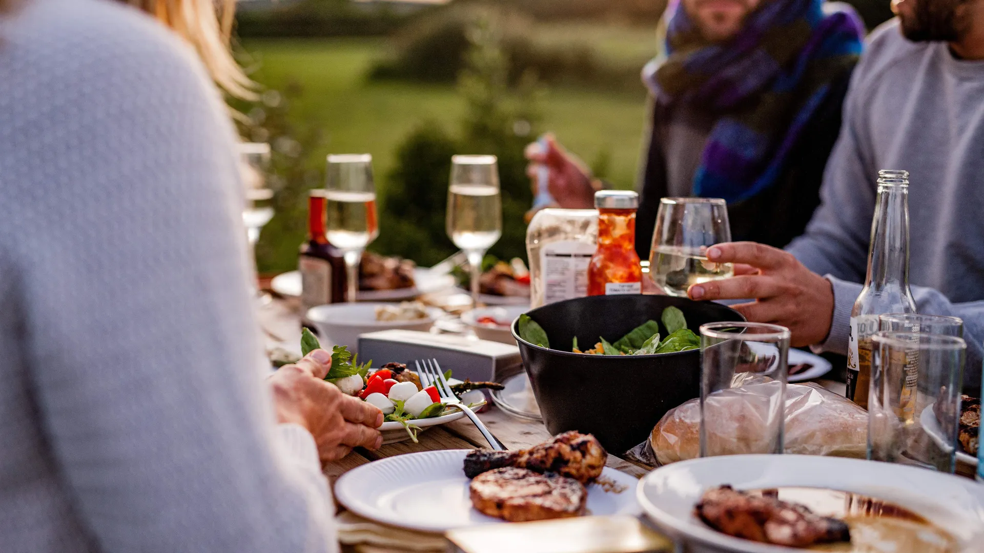 Friends sharing an outdoor meal at a table with wine glasses and plates of food at sunset