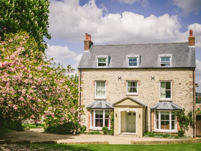 Stone farmhouse with grey slate roof and dormer windows, framed by pink blossom trees under a blue sky