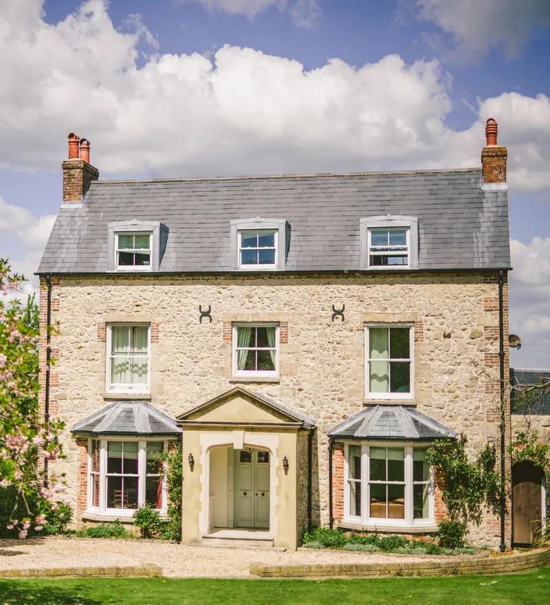 Stone farmhouse with grey slate roof and dormer windows, framed by pink blossom trees under a blue sky