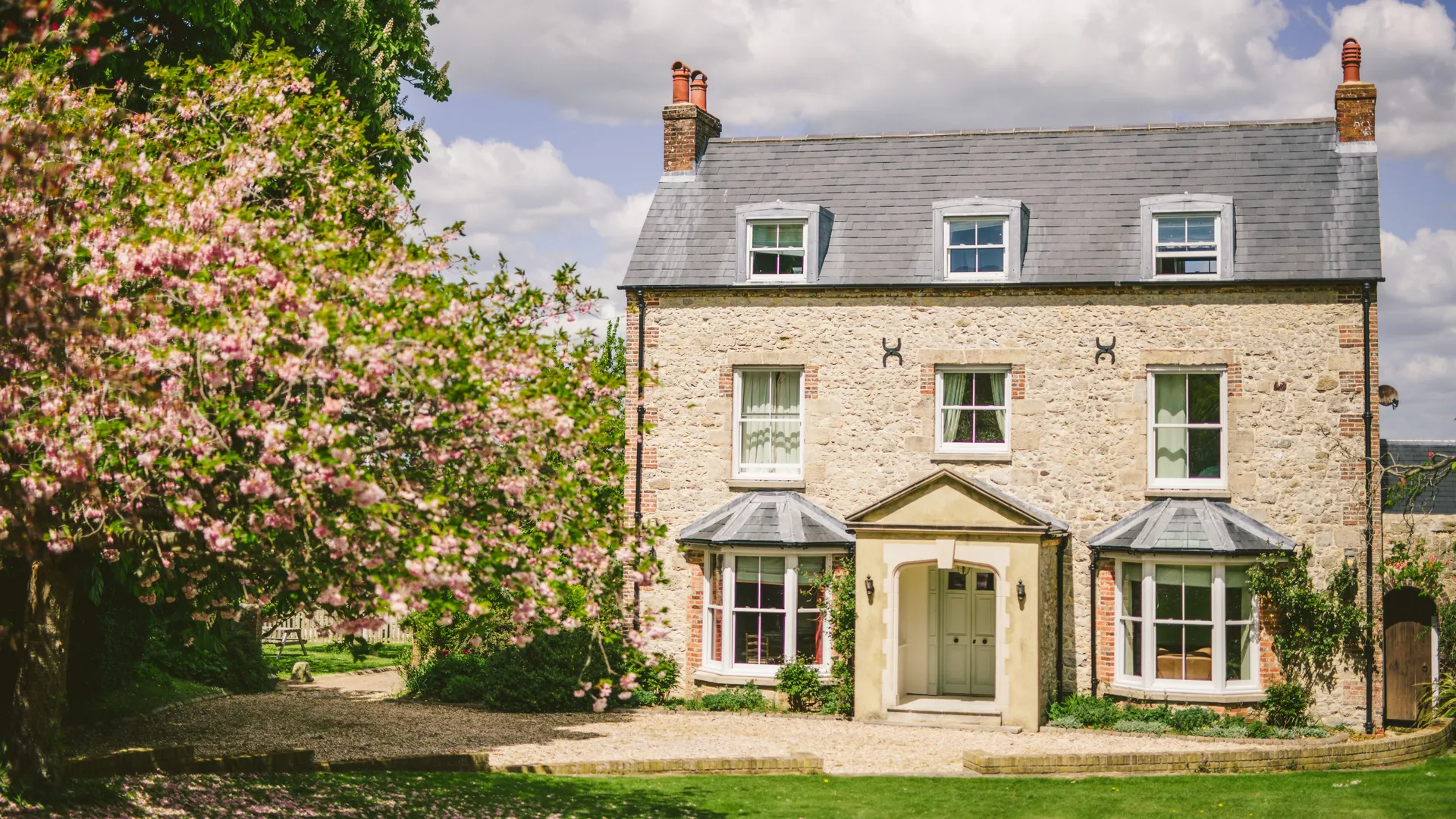 Stone farmhouse with grey slate roof and dormer windows, framed by pink blossom trees under a blue sky
