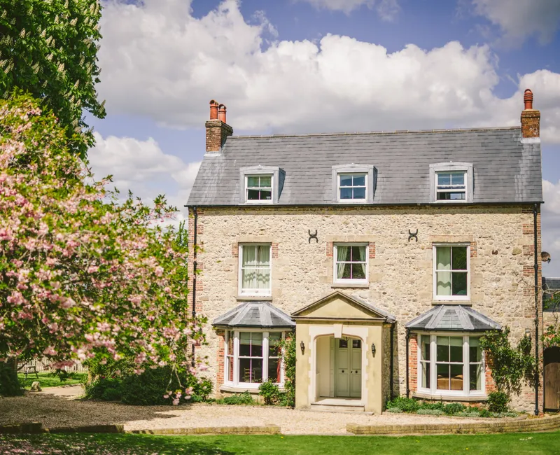 Stone farmhouse with grey slate roof and dormer windows, framed by pink blossom trees under a blue sky