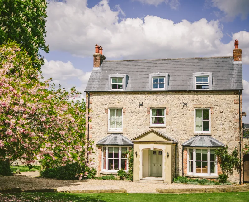 Stone farmhouse with grey slate roof and dormer windows, framed by pink blossom trees under a blue sky