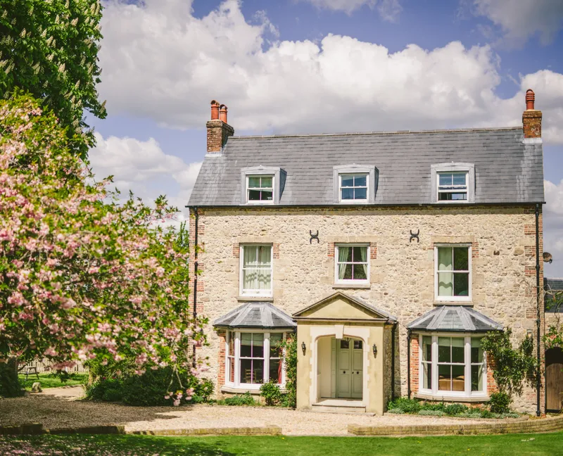 Stone farmhouse with grey slate roof and dormer windows, framed by pink blossom trees under a blue sky