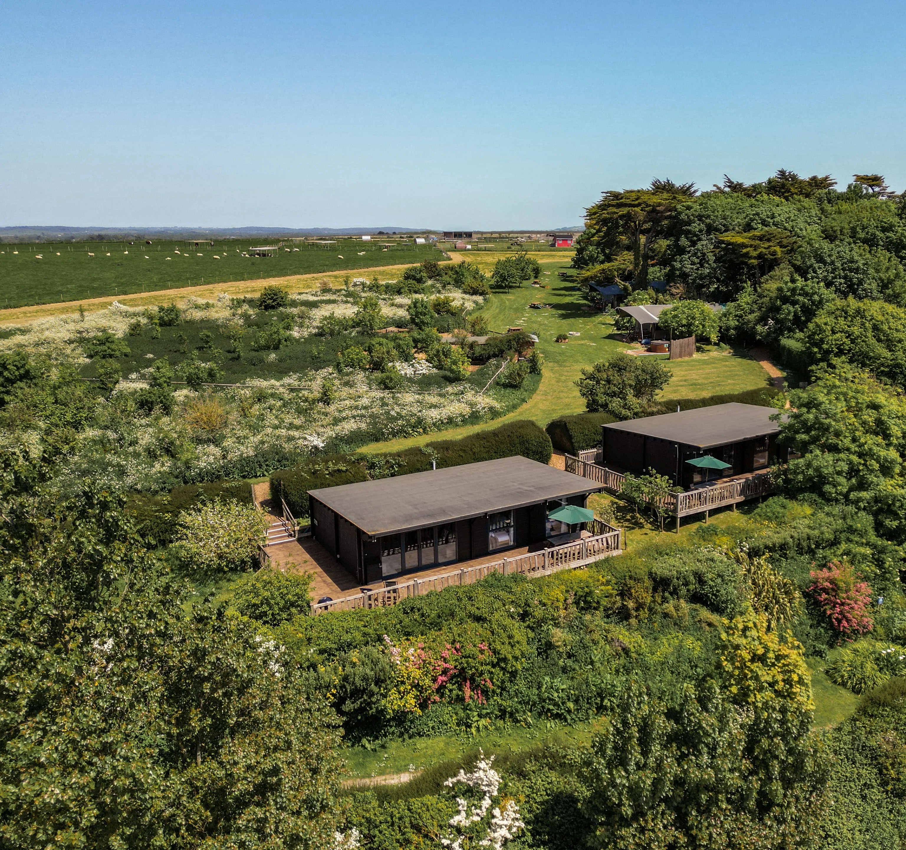 Aerial view of two modern lodges with wooden decks surrounded by lush gardens and countryside fields
