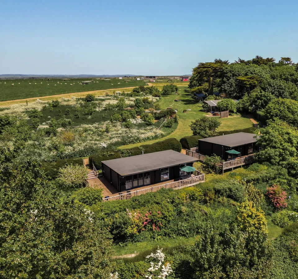 Aerial view of two modern lodges with wooden decks surrounded by lush gardens and countryside fields