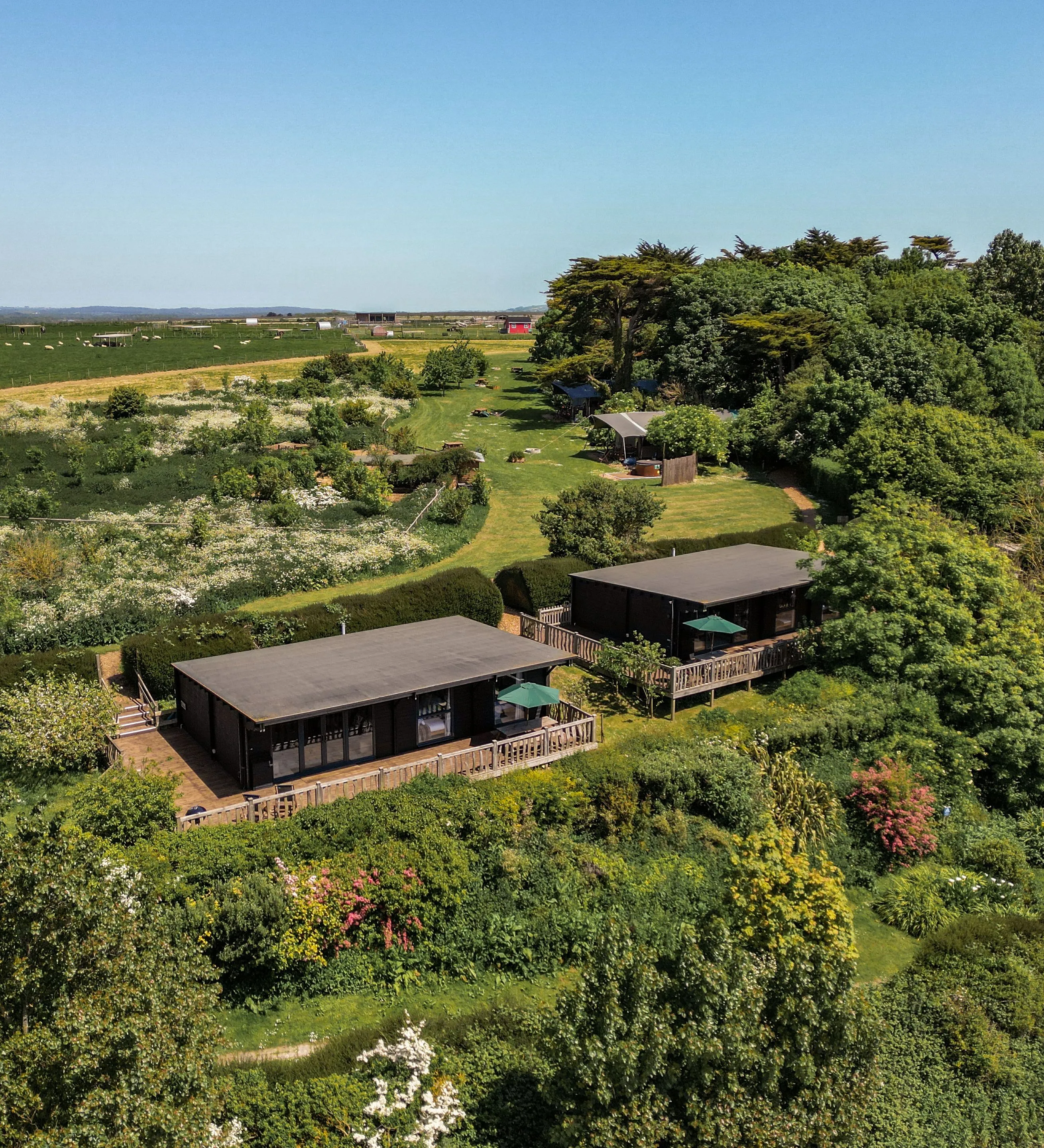 Aerial view of two modern lodges with wooden decks surrounded by lush gardens and countryside fields