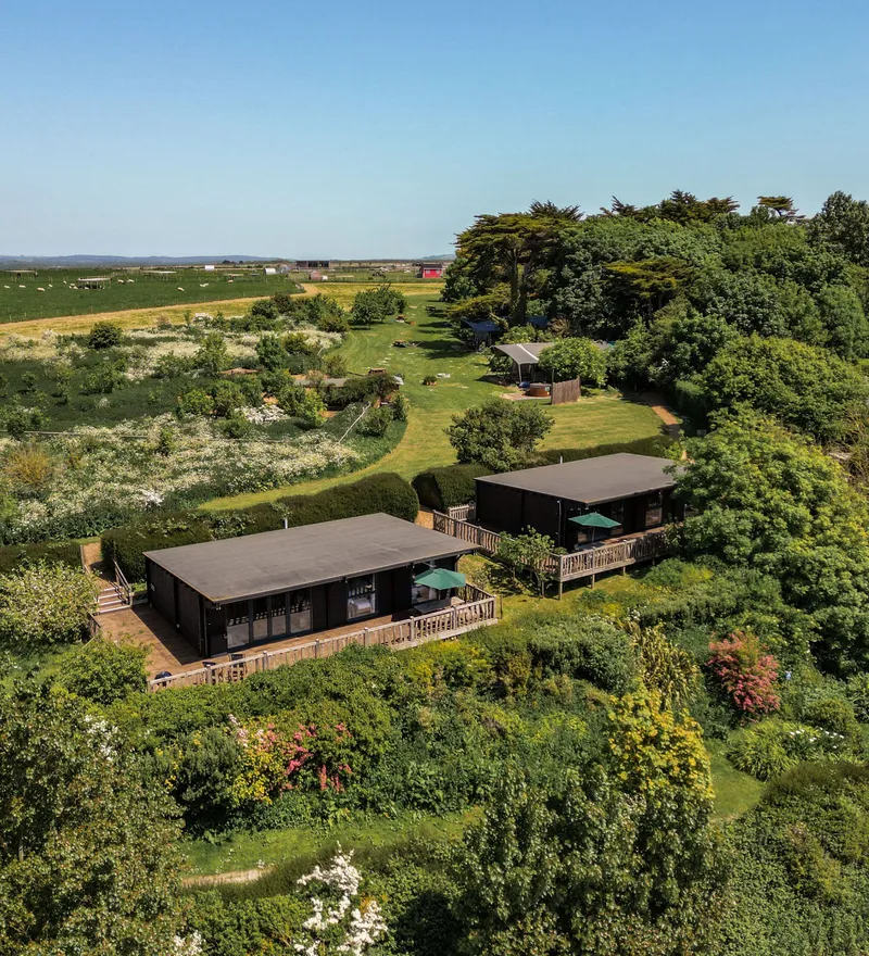 Aerial view of two modern lodges with wooden decks surrounded by lush gardens and countryside fields