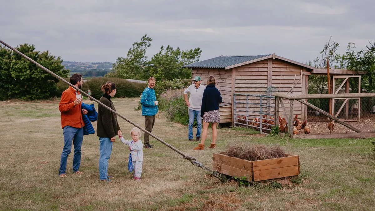 People chatting on a grassy farmyard beside a wooden chicken coop and fenced run