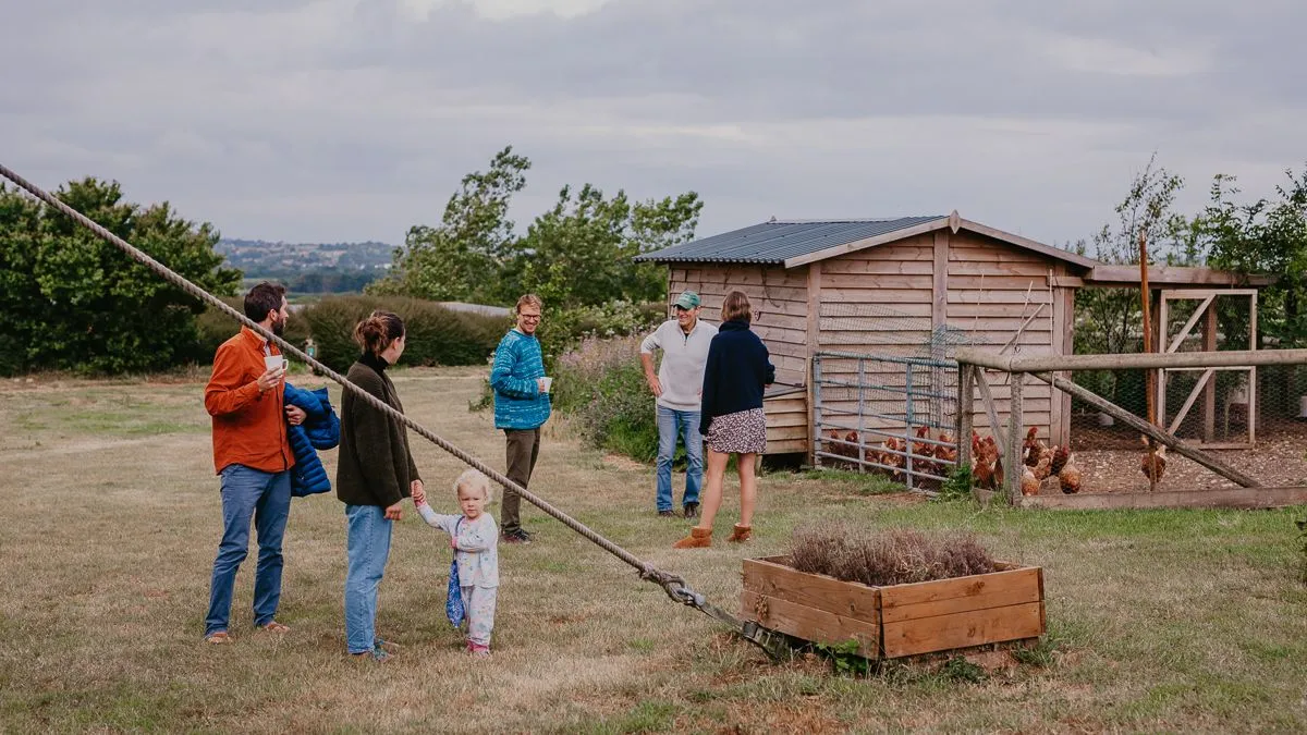 People chatting on a grassy farmyard beside a wooden chicken coop and fenced run