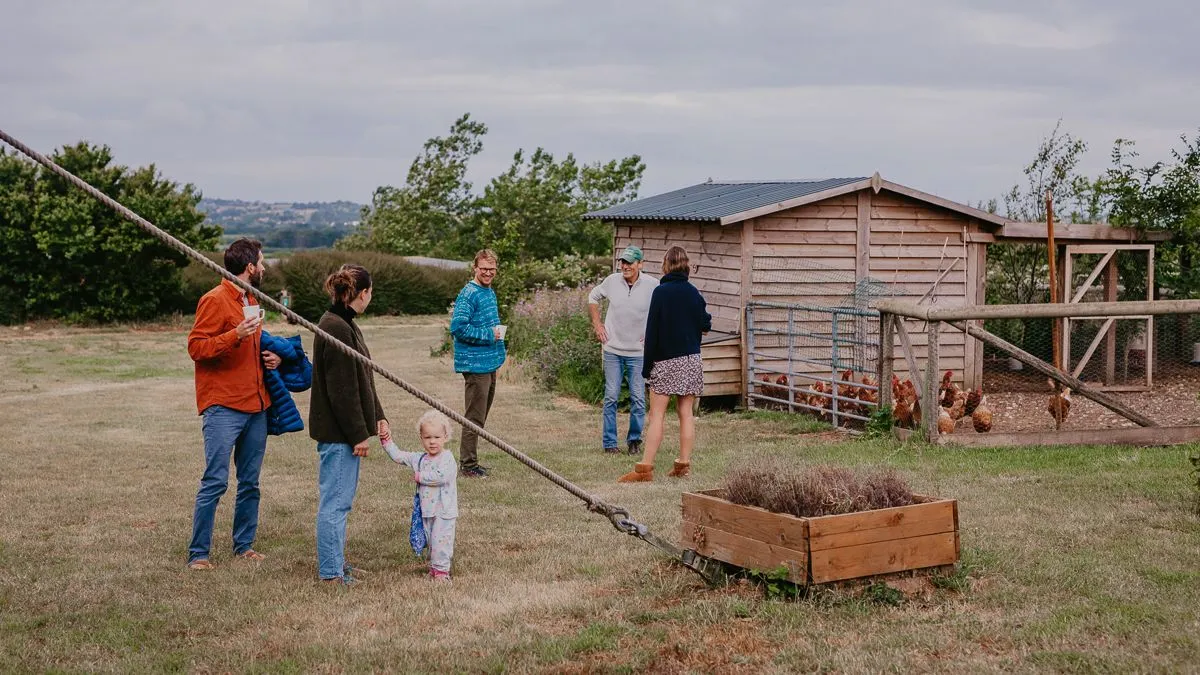 People chatting on a grassy farmyard beside a wooden chicken coop and fenced run
