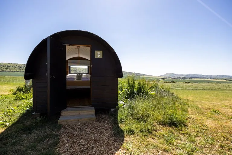 Small black camping pod with open door in a grassy field under a blue sky