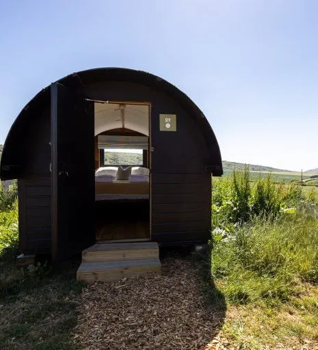 Small black camping pod with open door in a grassy field under a blue sky