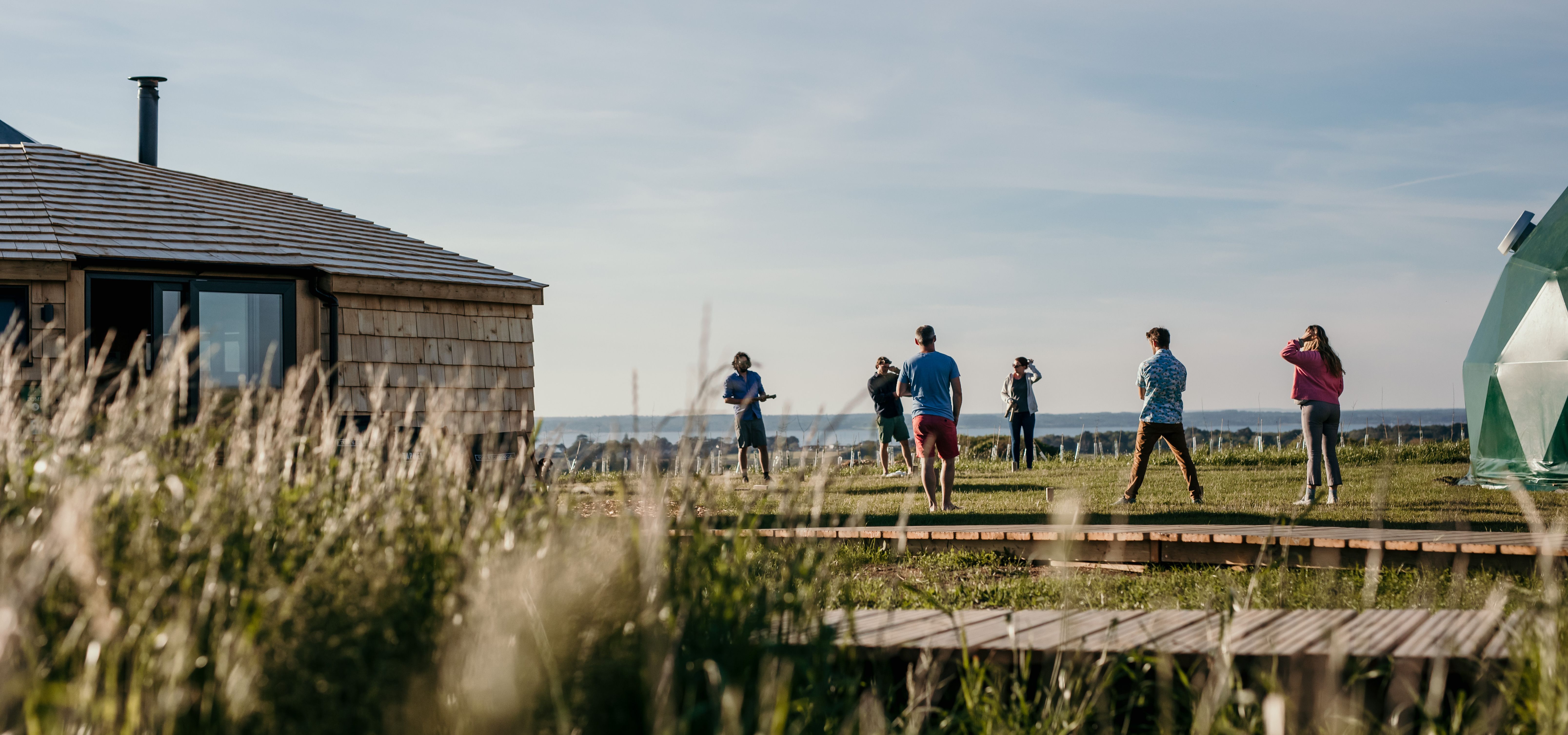 Guests standing on a grassy field beside a wooden lodge and a green geodesic dome at Tapnell Farm