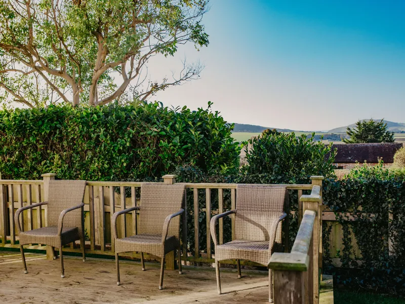 Three wicker chairs on a wooden deck with hedges and countryside views under a blue sky
