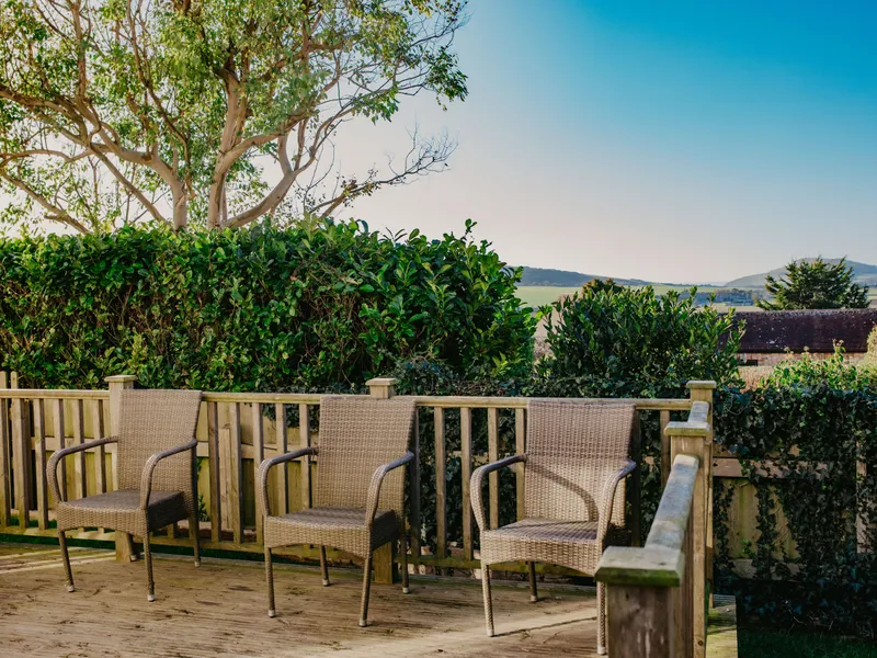 Three wicker chairs on a wooden deck with hedges and countryside views under a blue sky