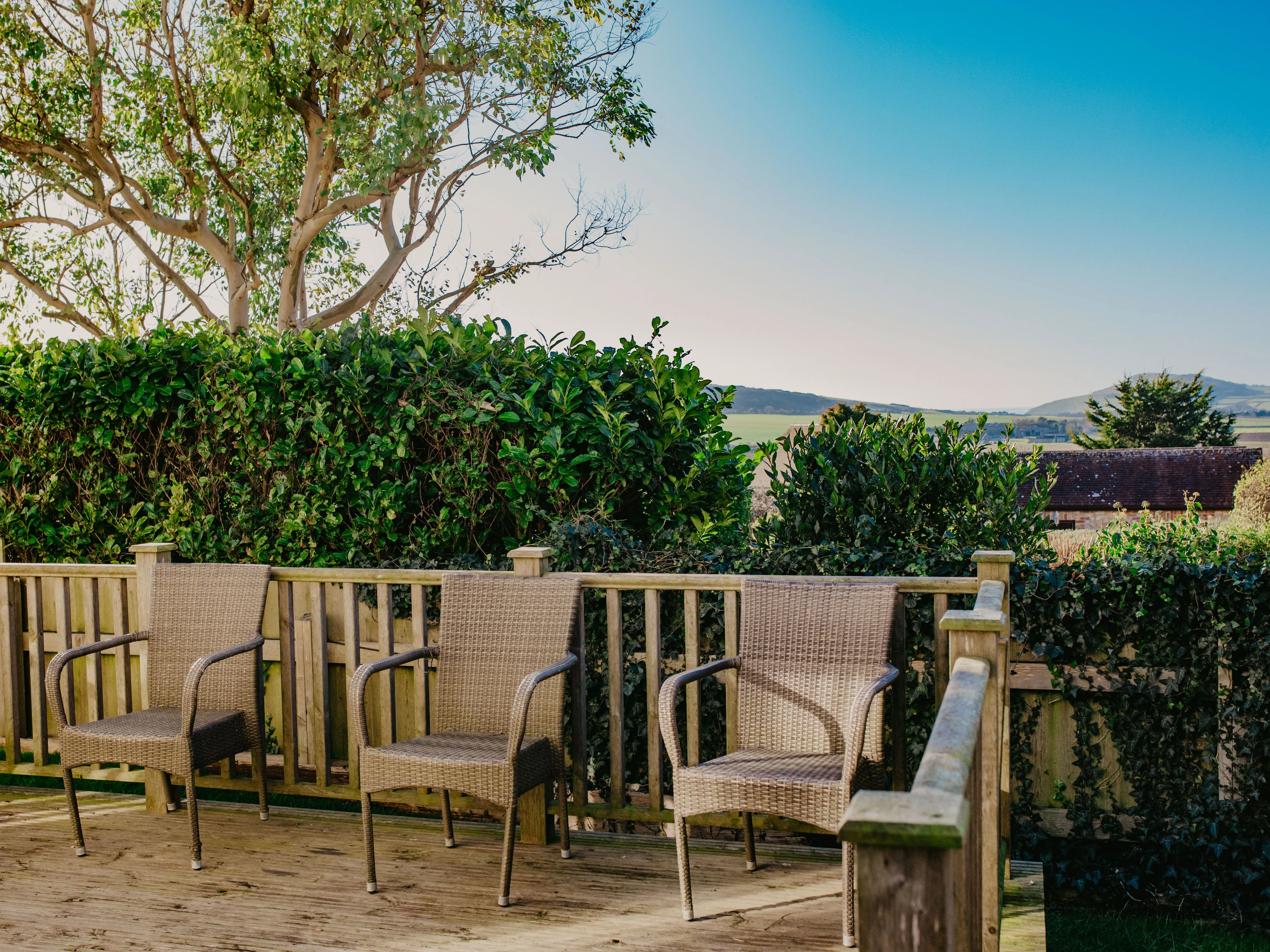 Three wicker chairs on a wooden deck with hedges and countryside views under a blue sky