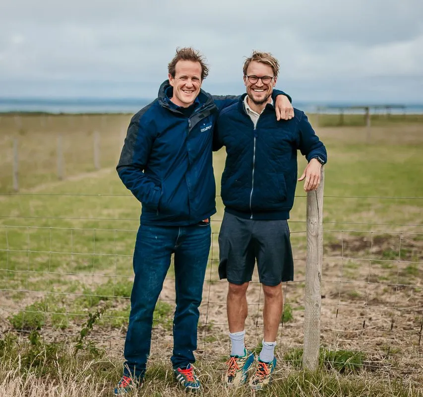 Two men standing by a wire fence in a grassy field at Tapnell Farm, smiling at the camera.