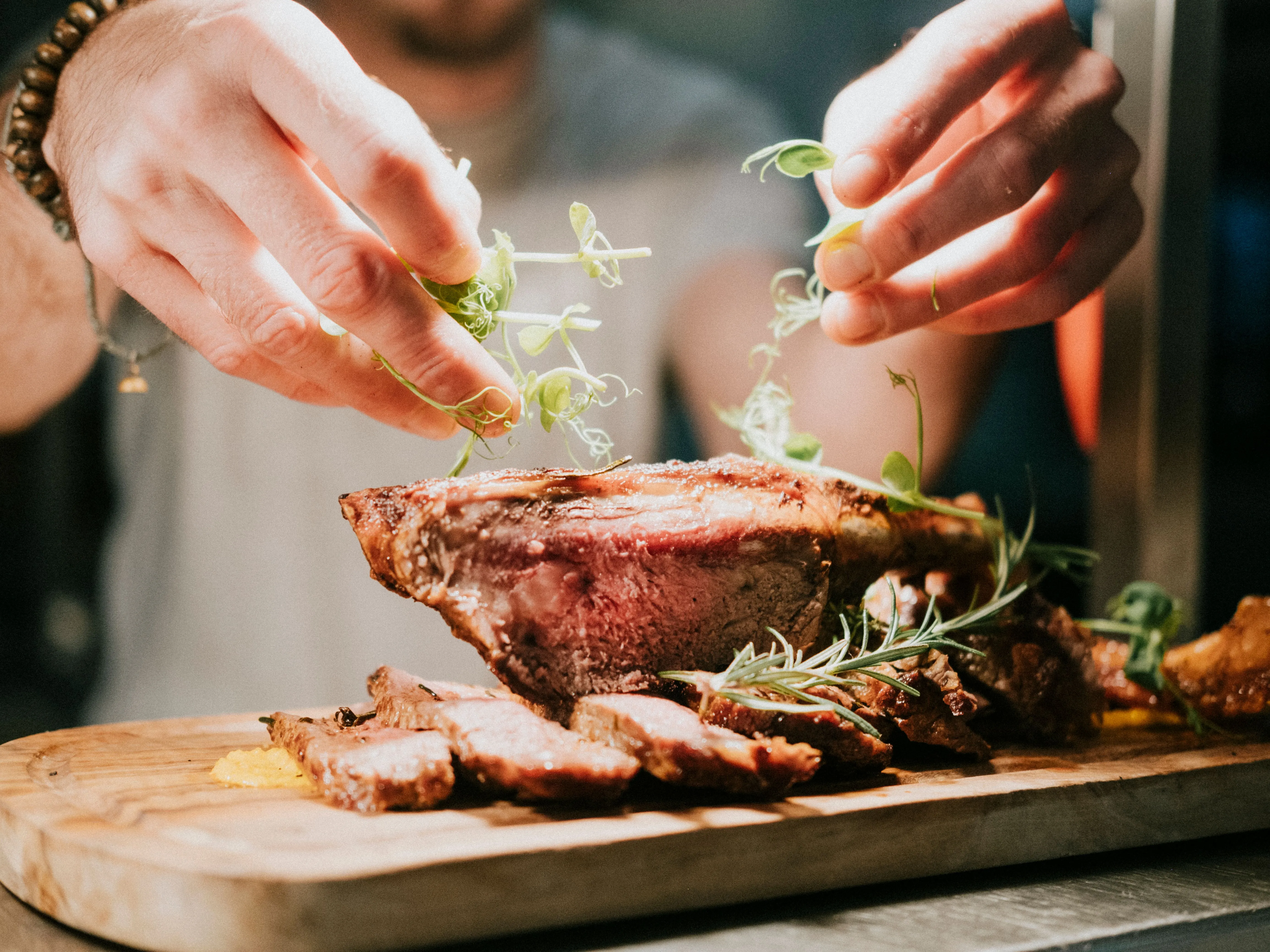 Hands garnishing sliced roast meat with microgreens on a wooden board