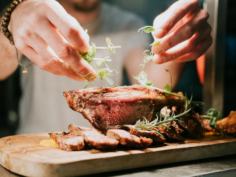 Hands garnishing sliced roast meat with microgreens on a wooden board