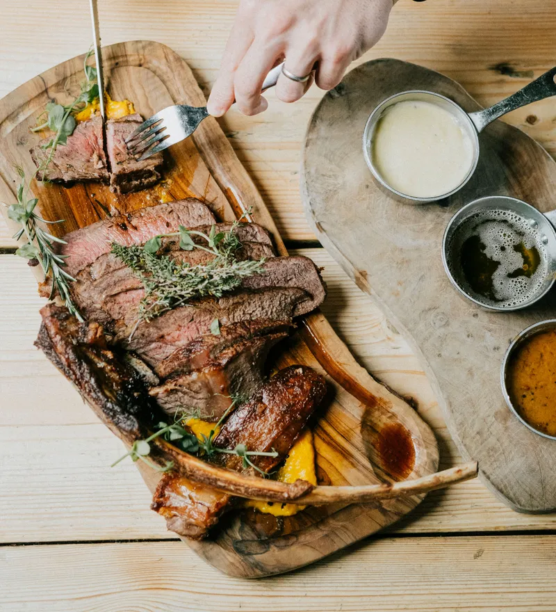 Hands slicing roast beef on a wooden board with herbs, beside small pans of gravy and sauce on a wooden table