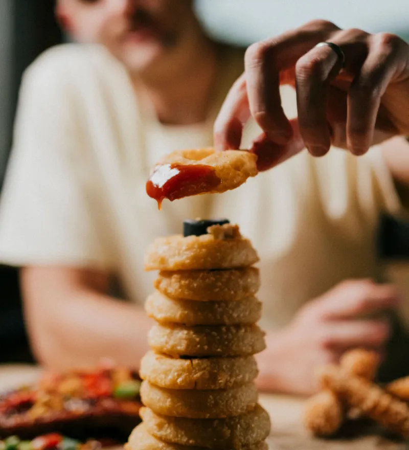Person dipping an onion ring into ketchup beside a tall stack of onion rings on a table