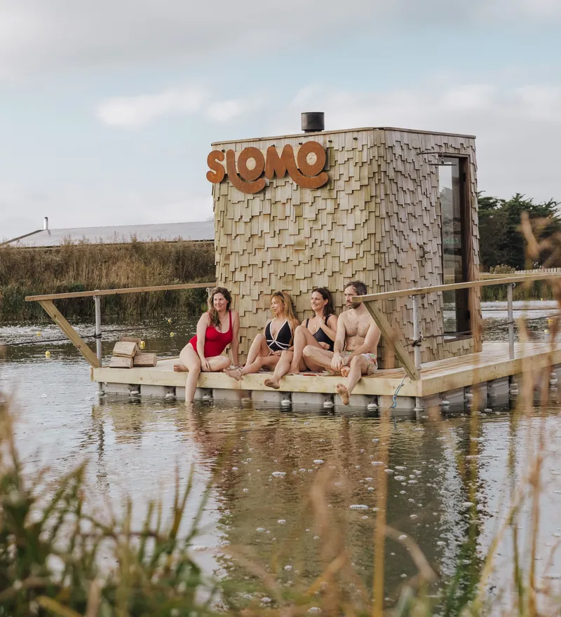 Four people in swimwear sitting on a wooden floating sauna platform beside a shingled hut with a large “SLOMO” sign, on a calm lake.