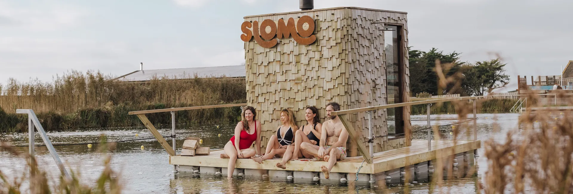 Four people in swimwear sitting on a wooden floating sauna platform beside a shingled hut with a large “SLOMO” sign, on a calm lake.