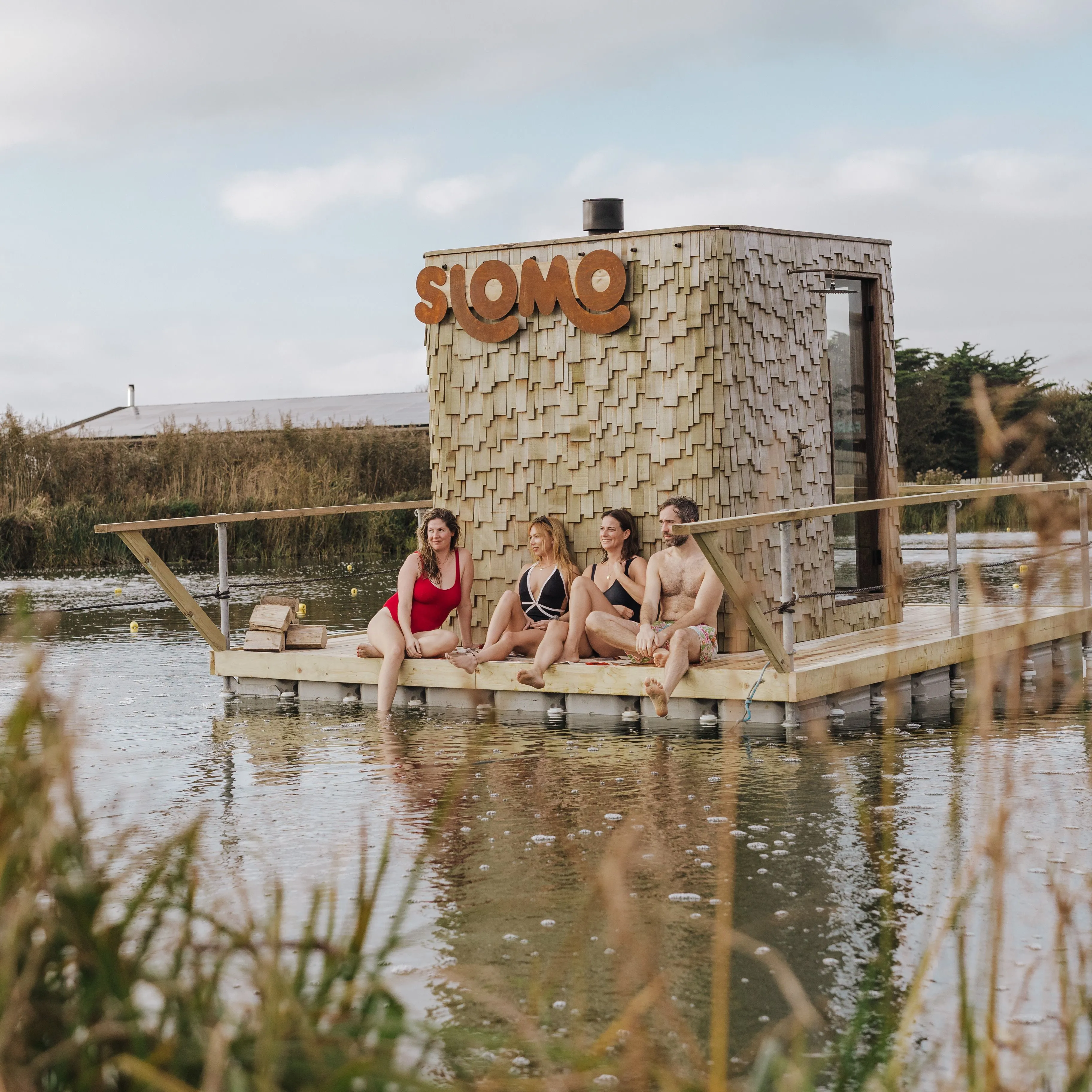 Four people in swimwear sitting on a wooden floating sauna platform beside a shingled hut with a large “SLOMO” sign, on a calm lake.