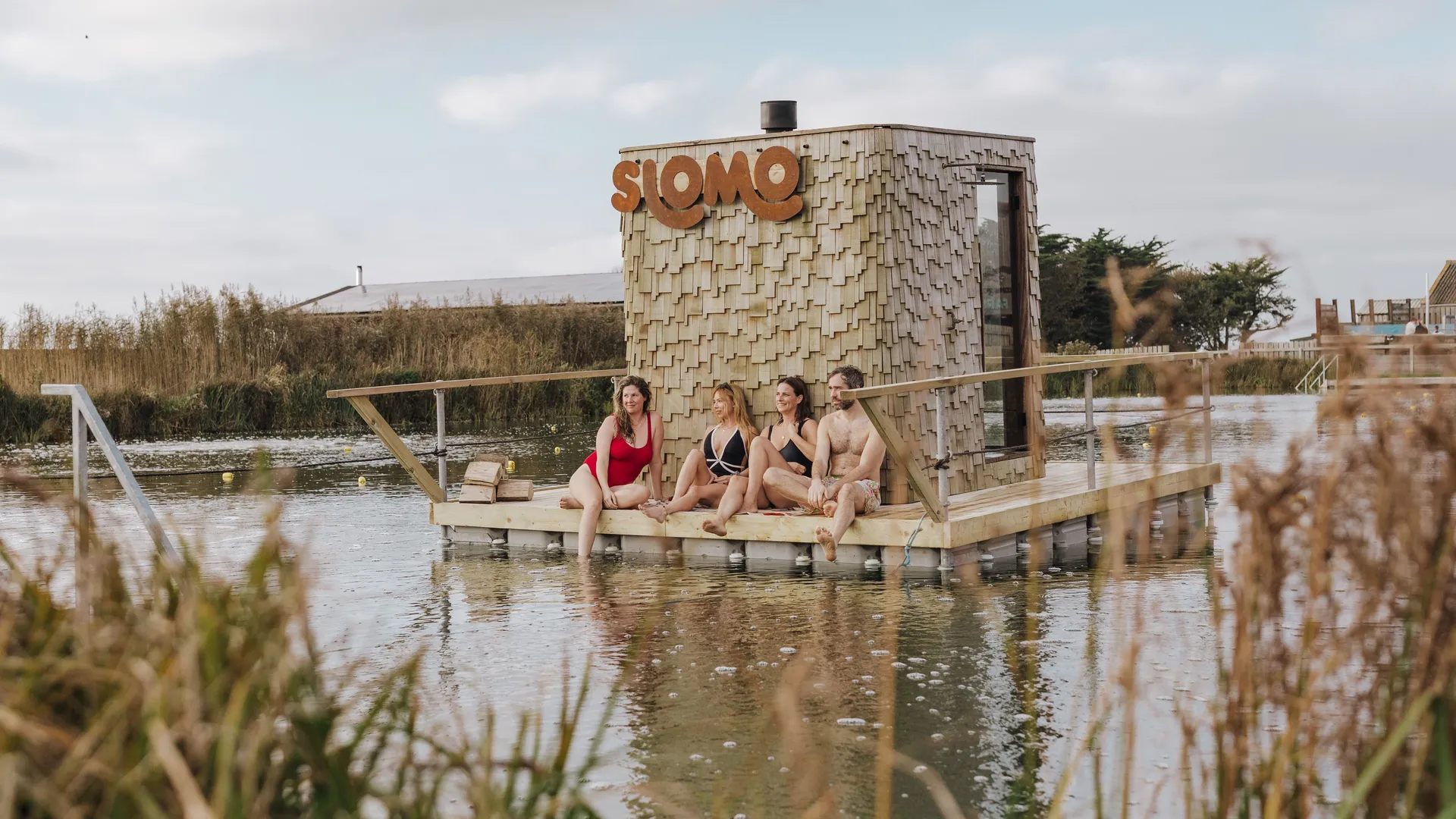 Four people in swimwear sitting on a wooden floating sauna platform beside a shingled hut with a large “SLOMO” sign, on a calm lake.
