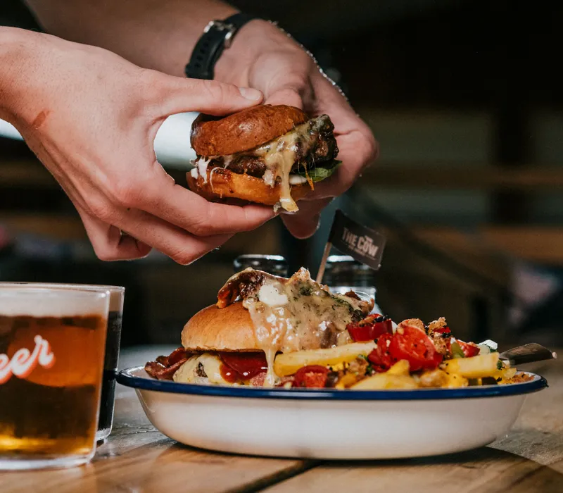 Hands holding a cheesy burger above a plate with another burger, chips and salad on a wooden table