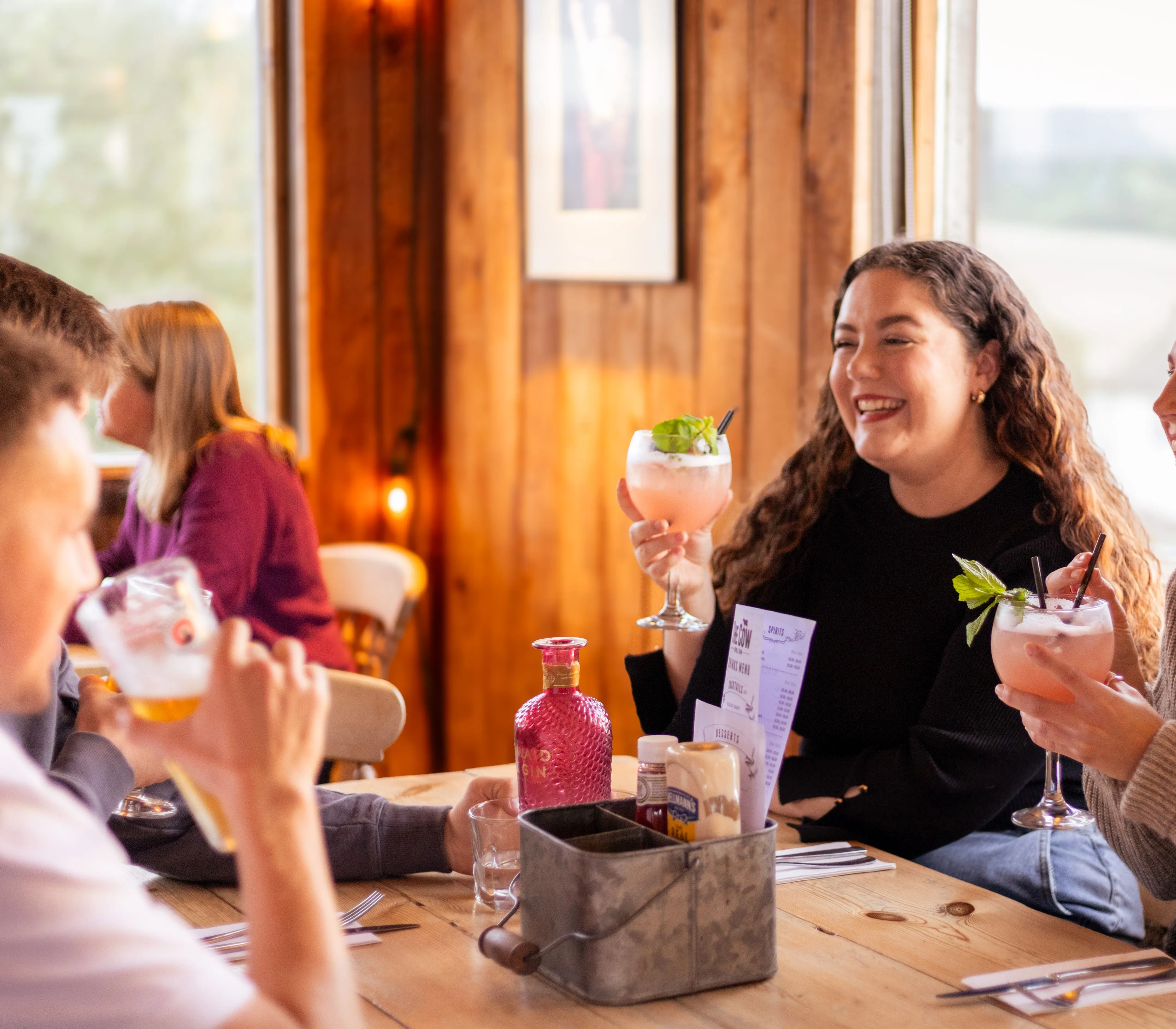 Friends laughing over cocktails at a rustic wooden table in a cosy farm restaurant