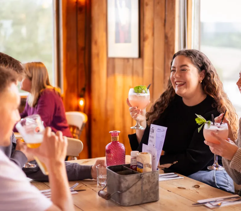 Friends laughing over cocktails at a rustic wooden table in a cosy farm restaurant